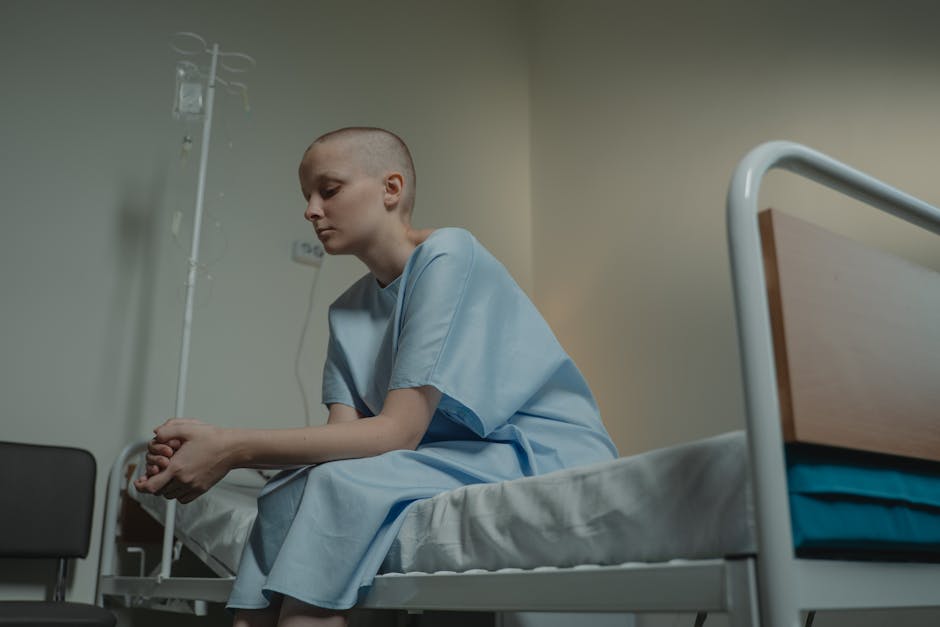 A person sits thoughtfully in a hospital room, wearing a hospital gown.